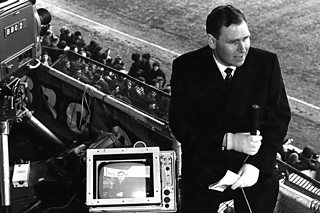 BBC presenter at pitchside with camera rig and monitor, crowd behind