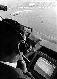 TV director in overhead booth watching monitor, pitch visible below